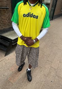 Zeleno-žlutý dres Adidas s černým nápisem, doplněný vzorovanými šortkami. Černé boty s bílými ponožkami, stojící na zpevněném povrchu.