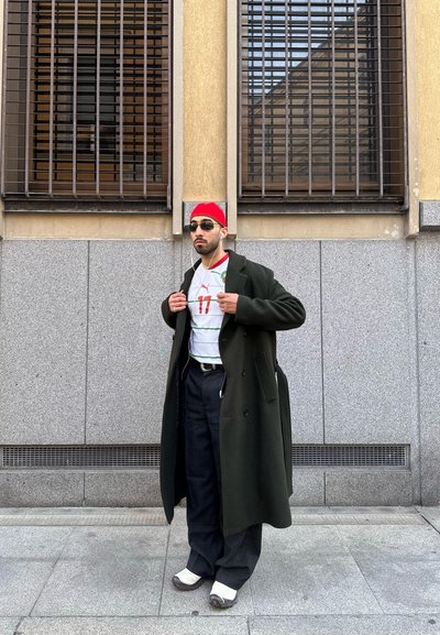 Hombre con abrigo negro, gorro rojo, gafas de sol, camiseta de fútbol blanca, pantalones oscuros y zapatillas blancas, de pie contra la pared beige de un edificio con ventanas con rejas.