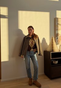 Femme debout à l'intérieur portant une veste à carreaux, un pull à losanges, un jean et des bottes marron, avec des arbres décoratifs sur un meuble sombre à côté.