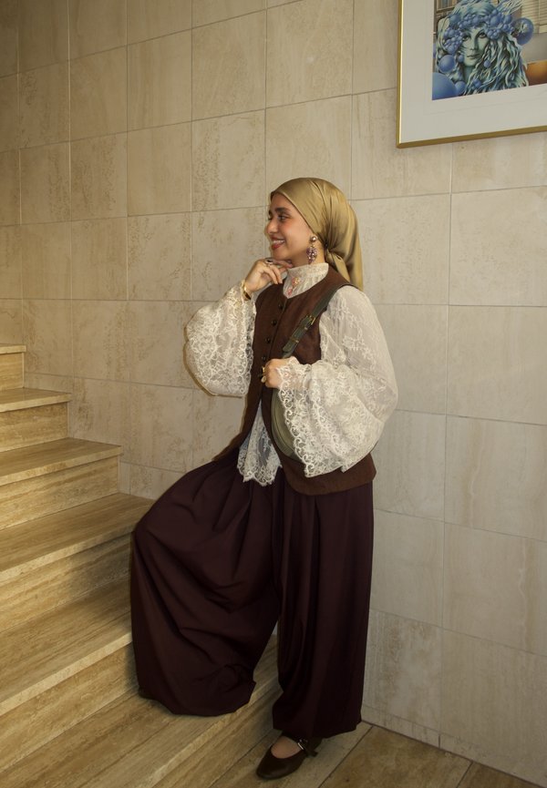 Aranyszínű hijabot viselő nő, fehér csipkefelsővel, barna mellénnyel és széles barna nadrággal, mosolyogva áll a lépcsőn, keze az állánál, a falon lévő keretezett festmény mellett.