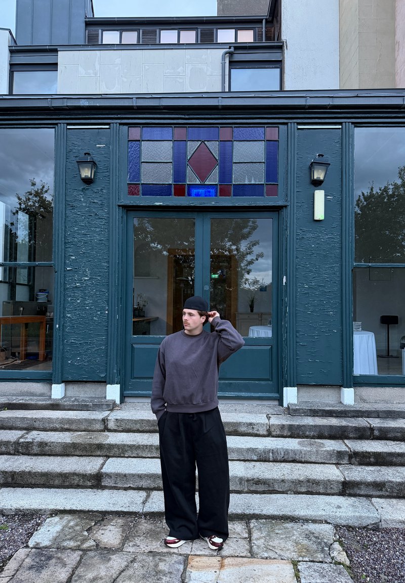 Uma pessoa jovem vestindo um boné, suéter e calças largas está em degraus de pedra em frente a um edifício verde com vitral acima das portas duplas.