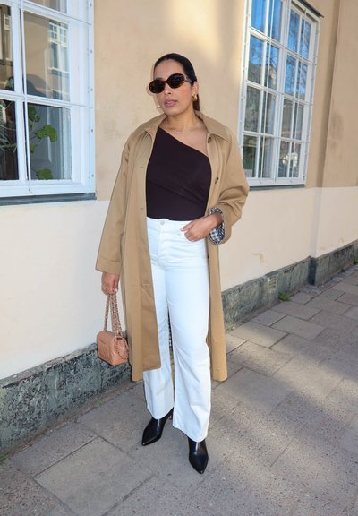 Persona con gafas de sol, abrigo camel, top negro de un hombro, pantalones blancos, botas negras, sosteniendo un bolso beige, de pie en una acera cerca de un edificio.