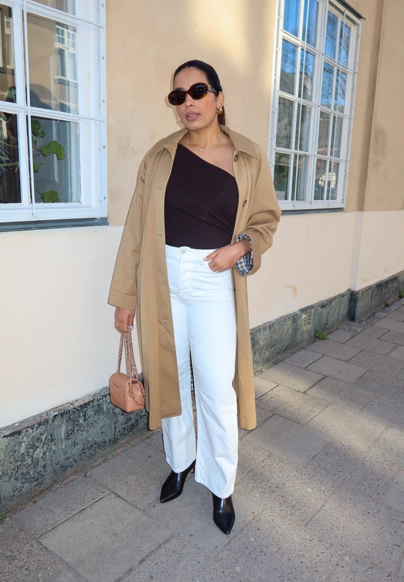 Personne portant des lunettes de soleil, un manteau couleur chameau, un top noir à une épaule, un pantalon blanc, des bottes noires, tenant un sac à main beige, debout sur un trottoir près d'un bâtiment.
