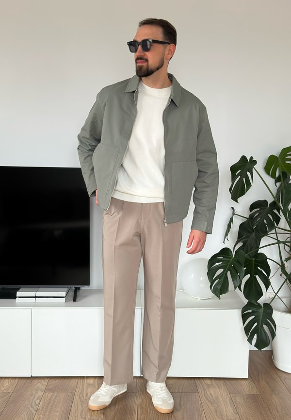 Jaqueta bomber cinza clara com fecho, suéter branco, calças bege e tênis bege. Óculos de sol pretos completam o visual. Ambiente interior.