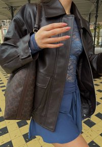 Femme portant une veste en cuir marron foncé et un haut en dentelle bleu, avec un sac à main marron à motifs, portant une jupe bleue sur un sol carrelé.