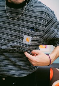 Homem a usar uma t-shirt riscada da Carhartt com óculos no bolso do peito, ajustando a camisa; usando um colar de corrente e uma pulseira trançada no pulso direito.