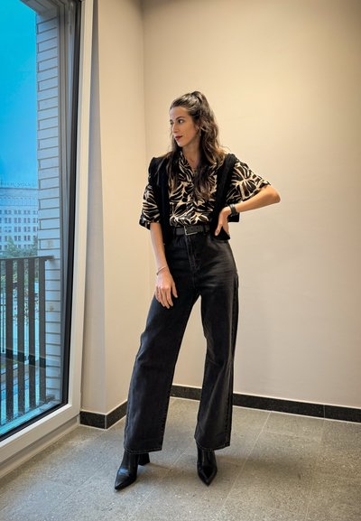 Jeans de tiro alto y pernera ancha negros, camisa estampada con un diseño abstracto en tonos beige y negro, chaleco negro ajustado y botas de tobillo de punta.