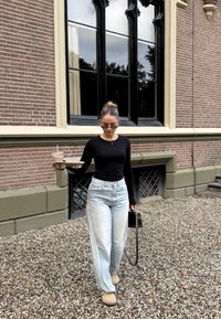 Langärmliges schwarzes Oberteil, hellblaue weit geschnittene Jeans mit seitlichen Streifen, beigefarbene Slipper, hält ein Getränk und ein Tablett mit Essen, schwarze Tasche.