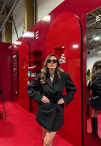 Robe blazer noire avec manches oversized ; modèle portant des lunettes de soleil foncées devant un fond rouge brillant avec des étagères de produits cosmétiques.