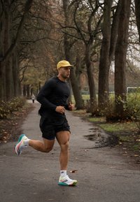 Bežec v čiernom tričku s dlhým rukávom a čiernych šortkách, s žltou šiltovkou a viacfarebnými bežeckými topánkami, behá po ceste obklopenej stromami.