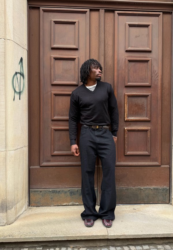 Homme avec des dreadlocks portant un pull noir et un pantalon large se tient devant une grande porte double en bois avec des graffitis sur un mur de pierre à proximité.