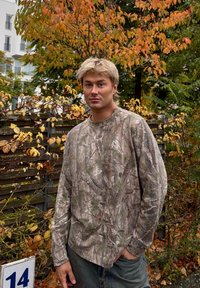 Camisa de larga de camuflaje, con un patrón de corteza de árbol en tonos terrosos, combinada con pantalones de mezclilla azul, contra un fondo de follaje otoñal.