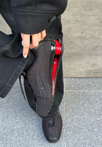 Schwarzer Sneaker mit Mesh-Obermaterial, roter sichtbarer Polsterung und schwarzer Sohle. Verfügt über ein Swoosh-Logo und Schnürsenkel, gehalten über einer grauen gekachelten Oberfläche.