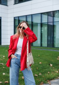 Κόκκινο oversized σακάκι, λευκό πουκάμισο, ανοιχτό μπλε φαρδιά τζιν και μπεζ τσάντα χιαστί με φράντζες. Γυαλιά ηλίου ολοκληρώνουν το λουκ.