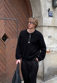Zwarte oversized sweater met een ronde hals, gedragen over zwarte losvallende broek. Een geweven zwarte tas wordt in één hand vastgehouden.