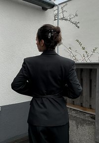 Blazer negro ajustado con un diseño entallado, que presenta una cintura ceñida y solapas anchas. La textura de la tela suave resalta los acentos minimalistas.
