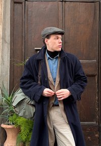 Jeune homme boutonnant son manteau, portant des vêtements superposés et une casquette plate, debout devant une grande porte en bois avec des plantes en pot à proximité.