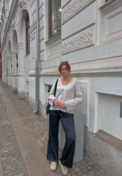 Modelo vistiendo un cárdigan gris de gran tamaño, blusa blanca y pantalones anchos negros. De pie contra una pared blanca texturizada, con una calle de adoquines.