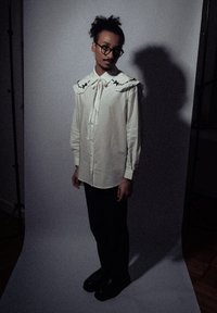 Chemise blanche à manches longues avec un grand col volanté comportant des motifs d'étoiles noires, associée à un pantalon noir et des chaussures noires imposantes.