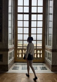 Femme en pull blanc et jupe debout près d'une grande fenêtre à carreaux, donnant sur un jardin avec une balustrade en pierre et des sentiers.