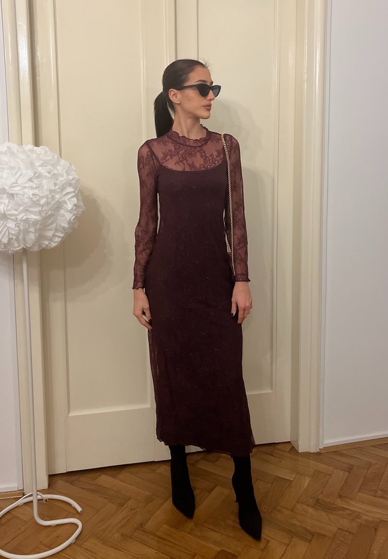 Femeie îmbrăcată într-o rochie lungă din dantelă neagră, cizme negre, ochelari de soare și o geantă cu lanț auriu, stând în interior lângă o ușă albă și o lampă înaltă albă.