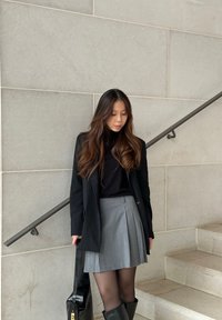 Μαύρο σακάκι πάνω από μαύρο πουλόβερ με ψηλό λαιμό, γκρι πλισέ φούστα και μαύρες μπότες μέχρι το γόνατο. Κρατώντας μια μαύρη τσάντα χειρός. Φωτεινό γκρι τοίχο από πέτρα στο φόντο.