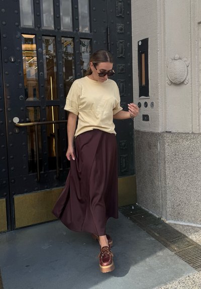 Mujer con gafas de sol vestida con una camiseta crema, una falda púrpura oscura y zapatos de plataforma marrones, de pie cerca de una puerta decorativa negra y una pared de piedra con un ornamento de tortuga.