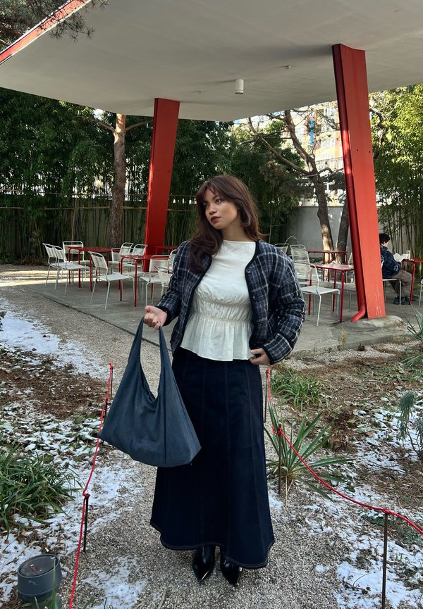 Mujer con chaqueta de cuadros y top blanco sosteniendo una bolsa grande azul, de pie en un camino cerca de asientos al aire libre de un café con pilares rojos.