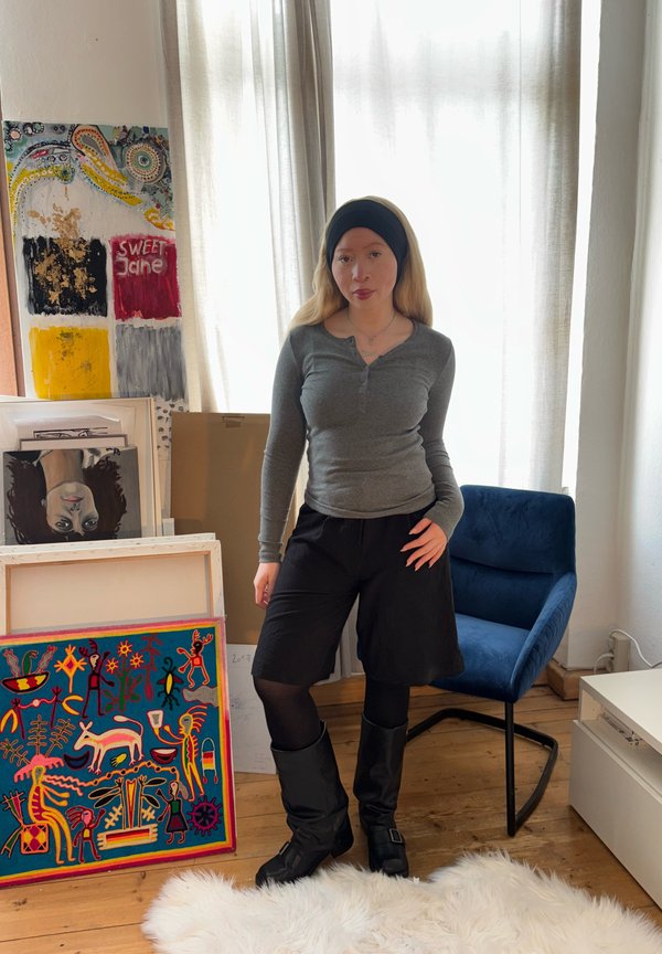 Grijze henley-shirt met lange mouwen, zwarte shorts en hoge zwarte laarzen. Kleurrijke kunstwerken met figuren en patronen zijn op de achtergrond te zien.