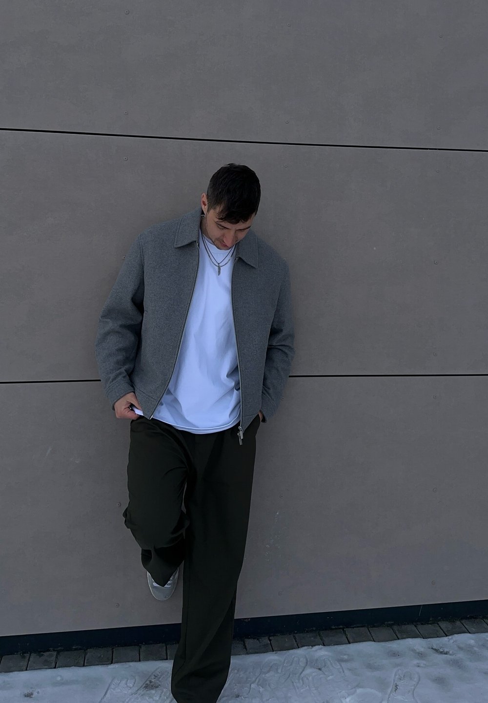 Veste grise zippée avec col, portée par-dessus un T-shirt blanc, associée à un pantalon vert foncé et des chaussures grises. Debout contre un mur clair.