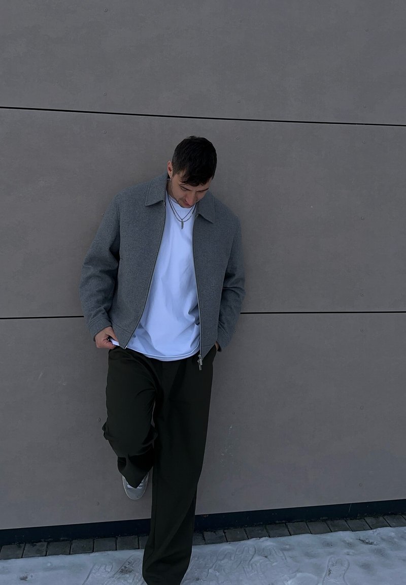 Veste grise à fermeture éclair avec col, portée sur un t-shirt blanc, associée à un pantalon vert foncé et des chaussures grises. Debout contre un mur clair.