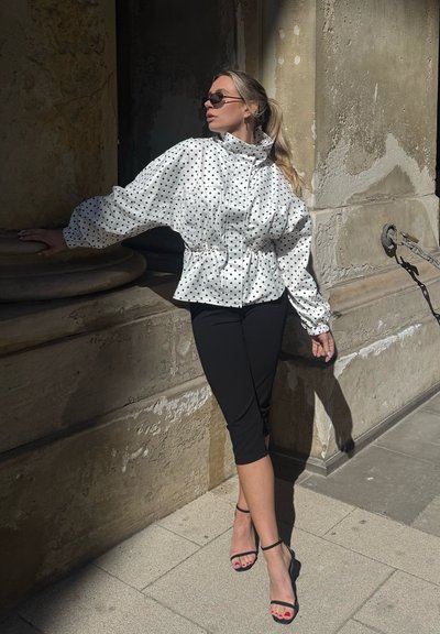 Mujer con pantalones cortos negros y chaqueta blanca de lunares se apoya en una pared de piedra, lleva gafas de sol y tacones negros con tiras bajo la luz del sol.