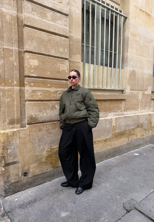 Personne portant des lunettes de soleil foncées, une veste aviateur vert olive et un pantalon noir ample, appuyée contre un mur en pierre beige sous une fenêtre avec barreaux.