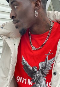 Red graphic t-shirt featuring a black-and-white angel design and the text "9N1M", worn under a light beige zip-up jacket. Accessorised with a silver chain necklace and hoop earrings.