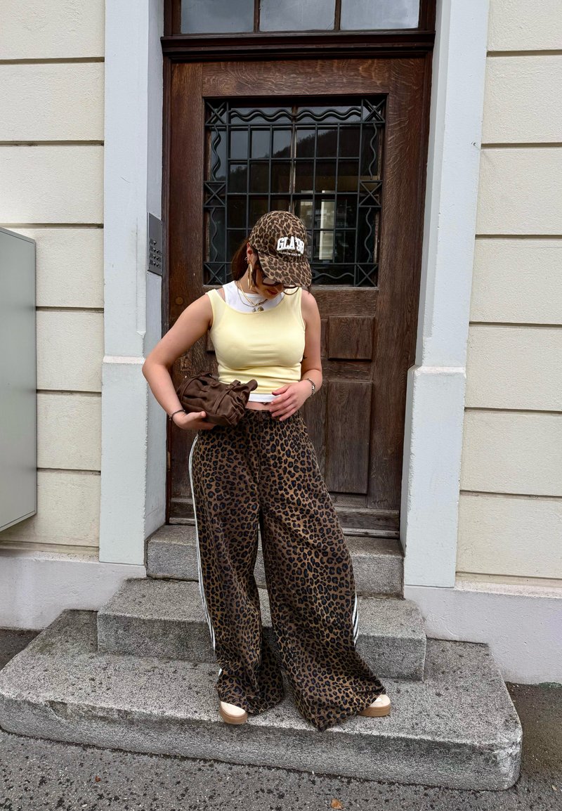 Vrouw draagt leopardprintbroek en pet, gele mouwloze top, houdt een bruine clutch vast, staat op de trap voor een houten deur.