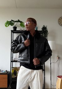 Veste bomber en cuir noir avec col en fourrure, dotée d'une fermeture éclair et d'une coupe courte. Portée sur une chemise noire et un pantalon crème.