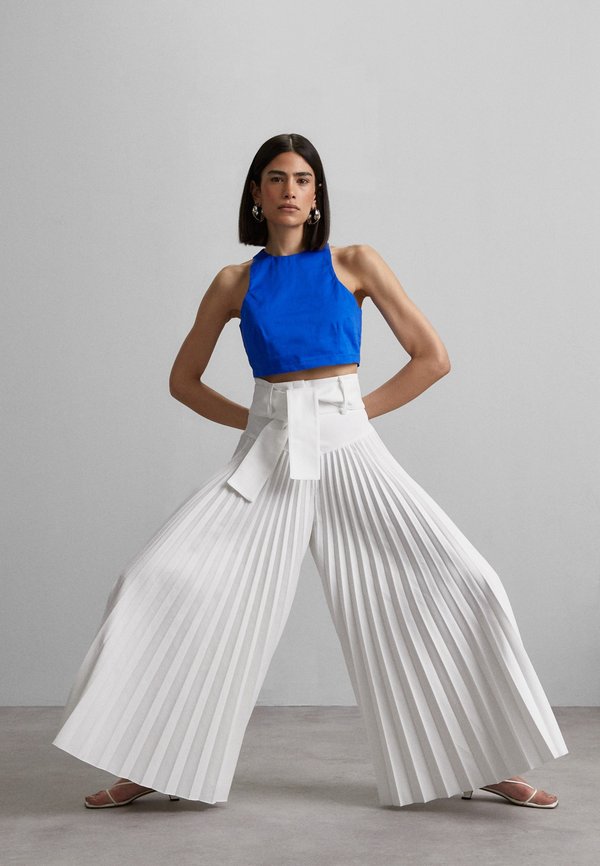 Κομμένο μπλε halter top σε συνδυασμό με φαρδιά, λευκά πλισέ παντελόνια που παρουσιάζουν μια μεγάλη λεπτομέρεια με ζώνη και διακριτική γυαλάδα. Το μοντέλο στέκεται με τα χέρια στους γοφούς.