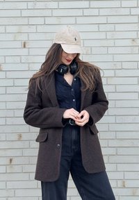 Jeune femme portant une casquette beige, un casque audio noir autour du cou, un blazer marron foncé, et un pantalon taille haute noir devant un mur de briques claires.