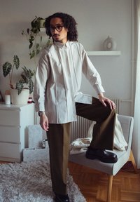 Lys grå skjorte med plisseret design, kontrasterende sømme; olivengrønne bukser; model poser med det ene ben oppe; sorte sko; indendørs miljø.