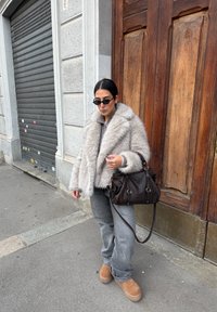 Világosszürke műszőrme kabát széles gallérral, sötétszürke farmernadrág, barna platform cipő és barna bőrtáska. A megjelenést napszemüveg teszi teljessé.