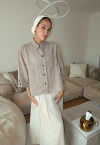 Svijetlosmeđa lanena košulja s kratkim rukavima, dva prednja džepa i tamnim gumbima, uparena s plisiranom suknjom u krem boji. Moderna unutrašnjost u pozadini.