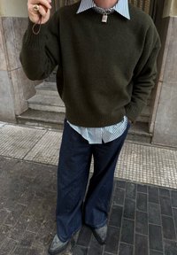 Personne portant un pull vert foncé sur une chemise rayée, un pantalon ample bleu foncé et des chaussures pointues, debout sur une rue carrelée près des marches d'un bâtiment.