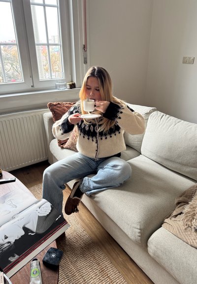 Mujer con suéter estampado sentada con las piernas cruzadas en el sofá, tomando té de una taza y platillo en una sala acogedora con un libro de fotos sobre la mesa.