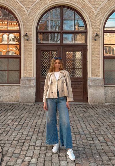 Joven con pelo ondulado que lleva una chaqueta beige, camisa blanca, vaqueros de pierna ancha y zapatillas blancas, de pie en una calle empedrada frente a una puerta arqueada.