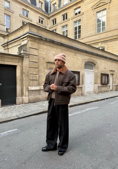 Hombre con chaqueta marrón, gorro de punto beige y pantalones anchos negros de pie en una calle de la ciudad con edificios históricos de piedra beige.