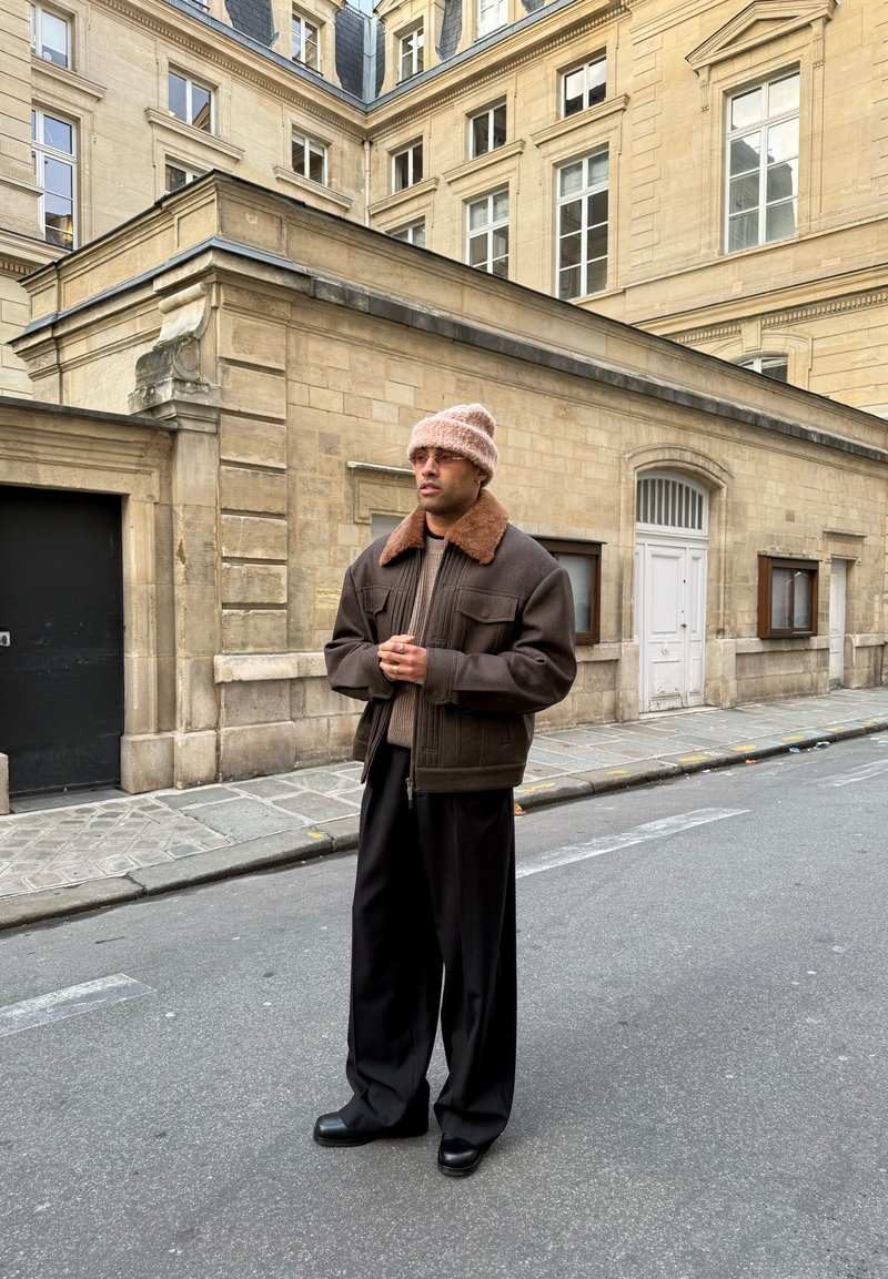 Homme portant une veste marron, un bonnet en tricot beige et un pantalon large noir, debout dans une rue de la ville bordée de bâtiments historiques en pierre beige.