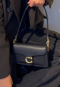 Czarna skórzana torebka z górnym uchwytem, o prostokątnym kształcie, ze złotymi ozdobami i klapą zamykaną na okrągły detal.