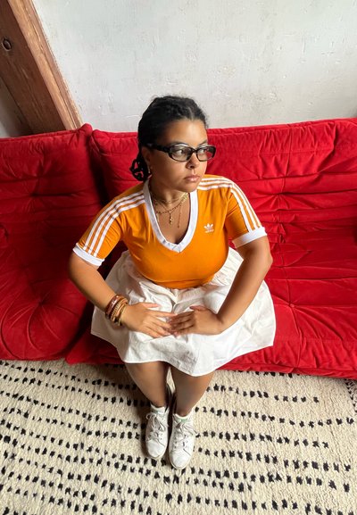 Camisa deportiva naranja con acentos blancos, falda blanca, pulseras en capas y zapatillas blancas, sentada en un sofá rojo con tejido texturizado.
