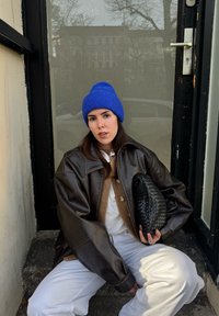 Jachetă maro din piele supradimensionat�ă, cămașă crem, pulover bej, pantaloni albi, căciulă albastră tricotată și o geantă clutch neagră țesută. În fundal se vede o ușă din sticlă.