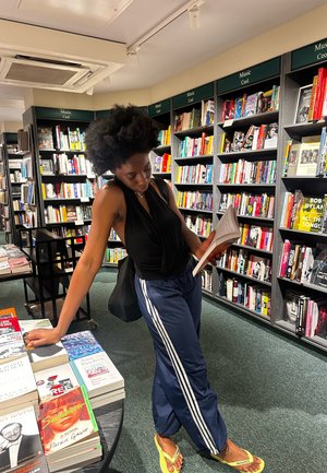 Top sin mangas negro, pantalones de chándal azul marino con rayas blancas, chanclas amarillas. Hojeando libros en un colorido ambiente de librería.